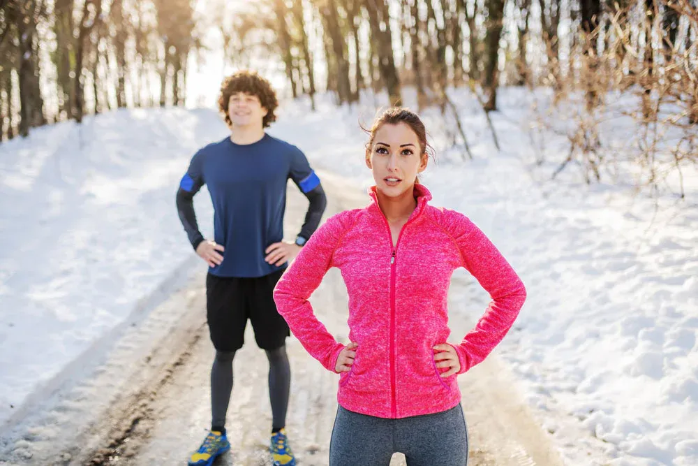 Two runners in winter running gear pausing on a snowy forest trail, showing weather-appropriate clothing
