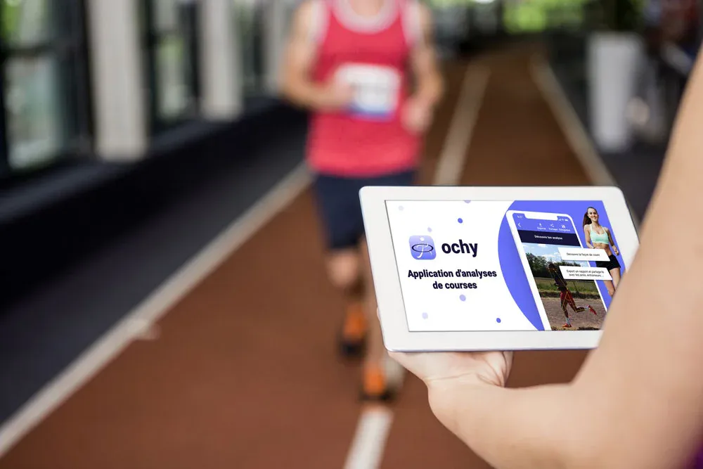 Tablet showing an early Ochy running analysis app screen while a runner jogs on an indoor track